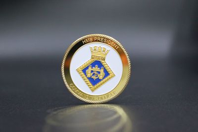 Gold-rimmed challenge coin with a blue-and-gold shield crest and white background