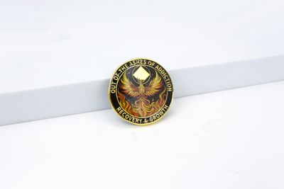Black-centered gold-tone coin showing a fiery phoenix with the wording Out of the Ashes of Addiction and Recovery & Growth