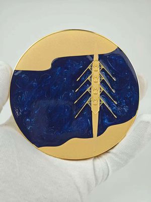 Large gold-tone disk with a blue textured water field and slender gold rowing crew in a long racing shell