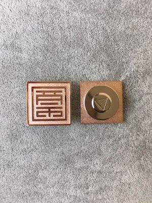 Front and back of copper-tone square lapel pin with raised seal-style linear motif and round clasp