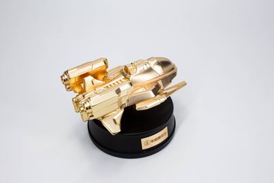 Gold-tone futuristic shuttle-style aircraft miniature with angular wings and prominent engines on black stand