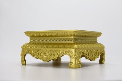 Gold-tone ornate rectangular elevated display stand with scrollwork relief and four short legs